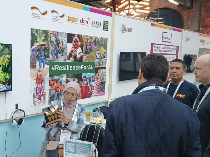 Personas observan una presentación en el stand conjunto de RED GIRDD-LAC y DiDRRN durante la Cumbre Global de Discapacidad 2025 en Berlín. En el fondo se aprecia un gran cartel con imágenes de personas con discapacidad participando en actividades comunitarias y el eslogan "#ResilienceForAll". Una mujer sostiene una tableta mostrando información, mientras una pantalla reproduce un video sobre reducción del riesgo de desastres inclusivo.